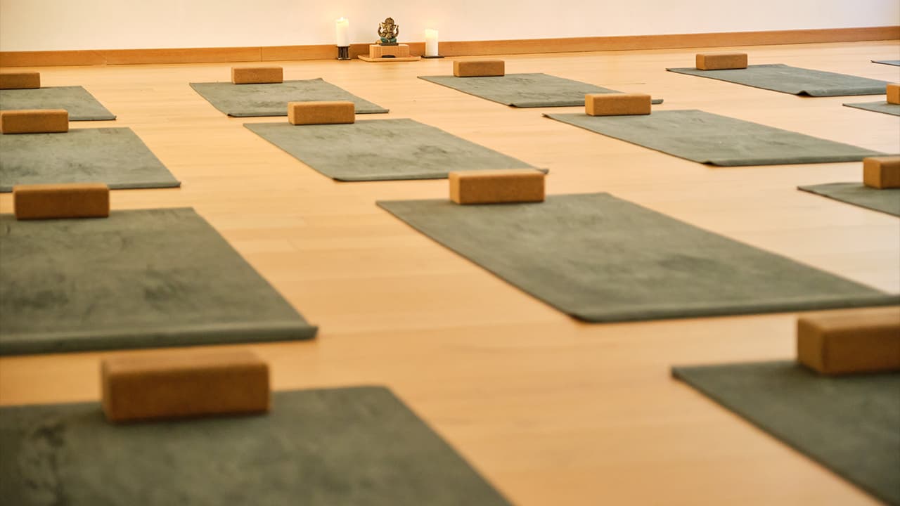 Yoga-Studio mit grauen Matten und Korkblöcken auf Holzboden, Ganesha-Statue und Kerzen im Hintergrund